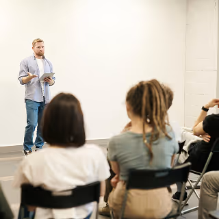 Man in casual clothes holding a tablet while giving a presentation to a seated group in a minimalist classroom setting.