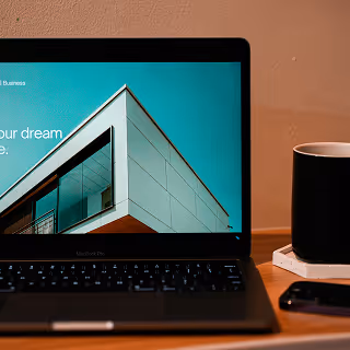 A black MacBook Pro on a desk displaying a sleek real estate website with the text “Build your dream home.” A black coffee mug sits beside it.