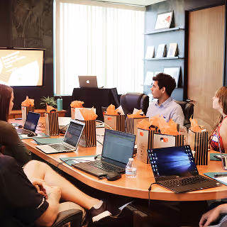 Group of professionals gathered in a corporate boardroom, looking at a presentation screen with laptops and branded gift bags on the table.