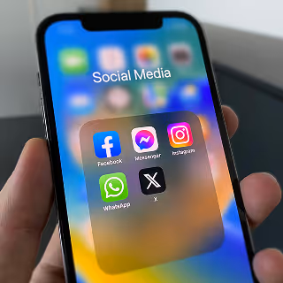 Close-up of a smartphone screen with Facebook, Messenger, Instagram, WhatsApp, and X apps in a “Social Media” folder, held in someone’s hand.