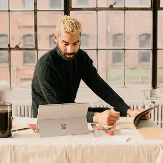 A man with bleached hair works at a table using a tablet and stylus, referencing printed materials while seated in a loft-style workspace with a large industrial window behind him.