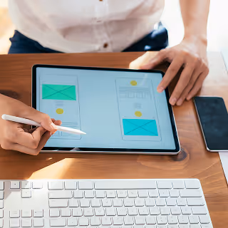 Close-up of a person using a stylus to design mobile wireframes on a tablet, placed next to a keyboard and smartphone on a wooden desk.