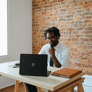 Professional man wearing glasses and a tie, working on a Microsoft Surface laptop at a wooden desk against a modern brick wall backdrop.