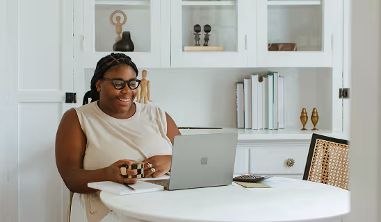 Woman smiling while working on a Microsoft Surface laptop at a round white table in a cozy, well-lit home office.