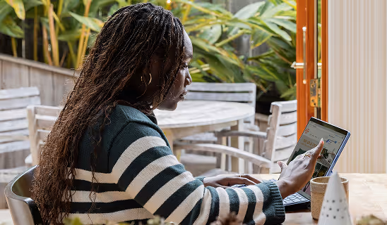 Focused woman in a striped sweater using a stylus to navigate a tablet while seated at an outdoor patio table surrounded by greenery.