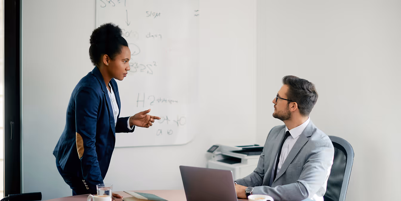 Businesswoman in a navy blazer pointing as she presents to a seated male colleague in a light grey suit, with a whiteboard in the background.