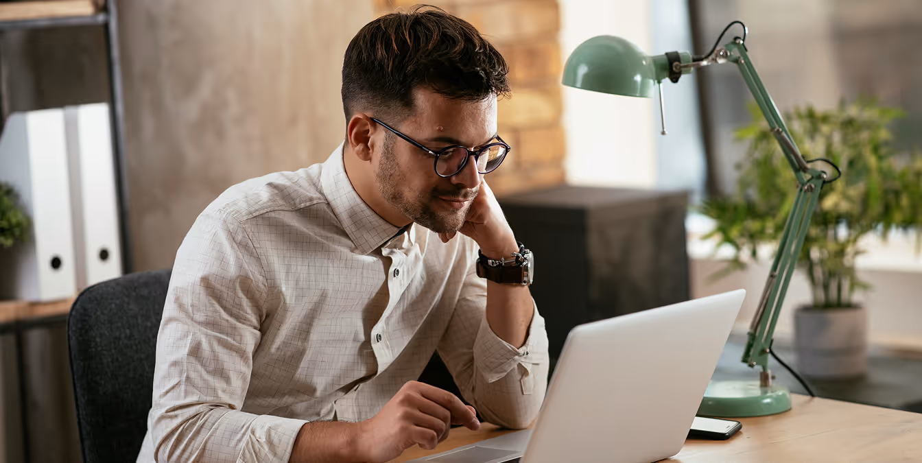 Focused man with glasses and a watch leaning over a laptop at a desk, surrounded by modern office decor including a green desk lamp and plants.