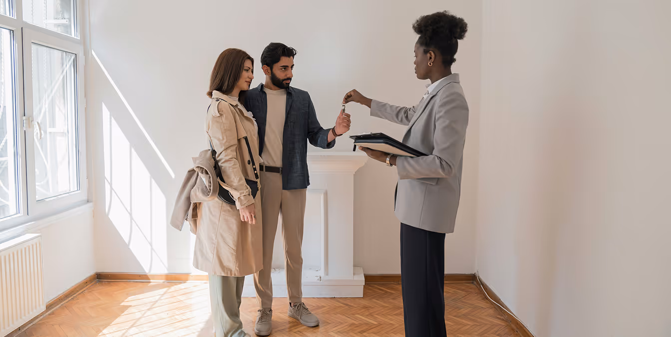 A real estate agent handing house keys to a young couple inside a new home, highlighting the personal touch in real estate transactions.