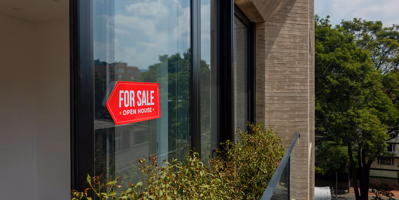 A red "For Sale - Open House" sign displayed on a modern glass balcony, overlooking a residential neighborhood.