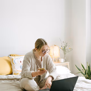 Woman sitting on a bed using a laptop and smartphone, working remotely from a cozy home environment.