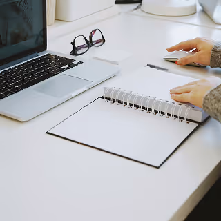 A professional working at a white desk with a laptop, notebook, and glasses, suggesting strategic planning or brand documentation.