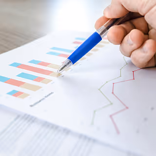 A close-up of a hand holding a pen over a chart with colorful bar graphs and trend lines, illustrating brand performance analysis.