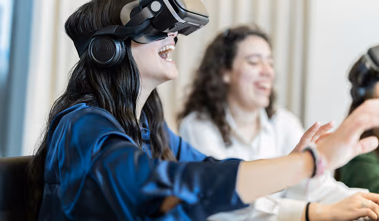 Smiling woman in a dark blue blouse wearing a VR headset, extending her hand forward while another woman laughs beside her, illustrating joy and social connection in virtual reality.