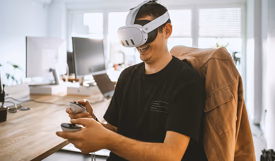 Man in a casual black shirt using hand controllers with a VR headset in a home office setting, emphasizing individual engagement in immersive tech.