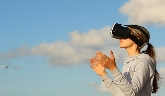 Woman outdoors wearing a VR headset, with her hands raised and a serene sky behind her, capturing exploration and wonder in virtual outdoor settings.