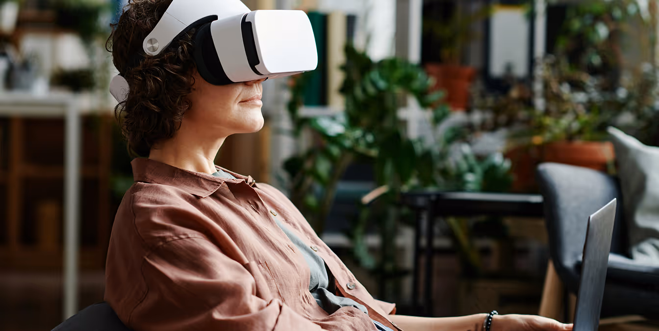 Woman sitting calmly on a sofa, wearing a VR headset while holding a laptop, surrounded by plants in a cozy modern room, symbolizing relaxed and immersive digital experiences at home.