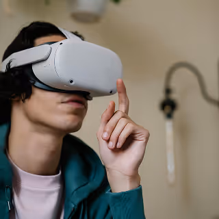 A young man wearing a white virtual reality headset and green hoodie sits indoors, gesturing with his index finger raised while immersed in a VR experience.