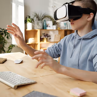 A teenager wearing a VR headset in a light-filled home office reaches forward with both hands, engaging with a virtual environment at a desk with a keyboard and mouse.