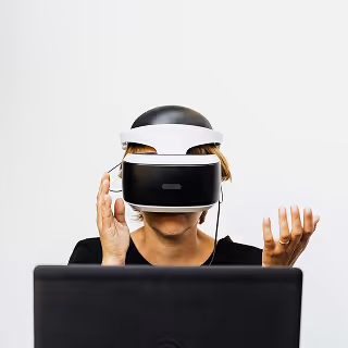 A woman wearing a black-and-white VR headset interacts with a virtual space while sitting in front of a laptop, her hands raised as if responding to stimuli.