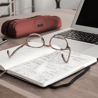Eyeglasses rest on an open notebook filled with handwritten notes beside a laptop and a red pencil case, suggesting content planning or budgeting.