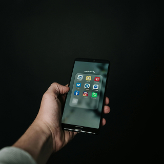 A person holds a smartphone with social media app icons displayed on the screen, against a dark, moody background.