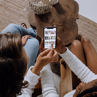 Overhead view of two women sitting by a wooden table, one pointing at a smartphone screen displaying a grid of images.