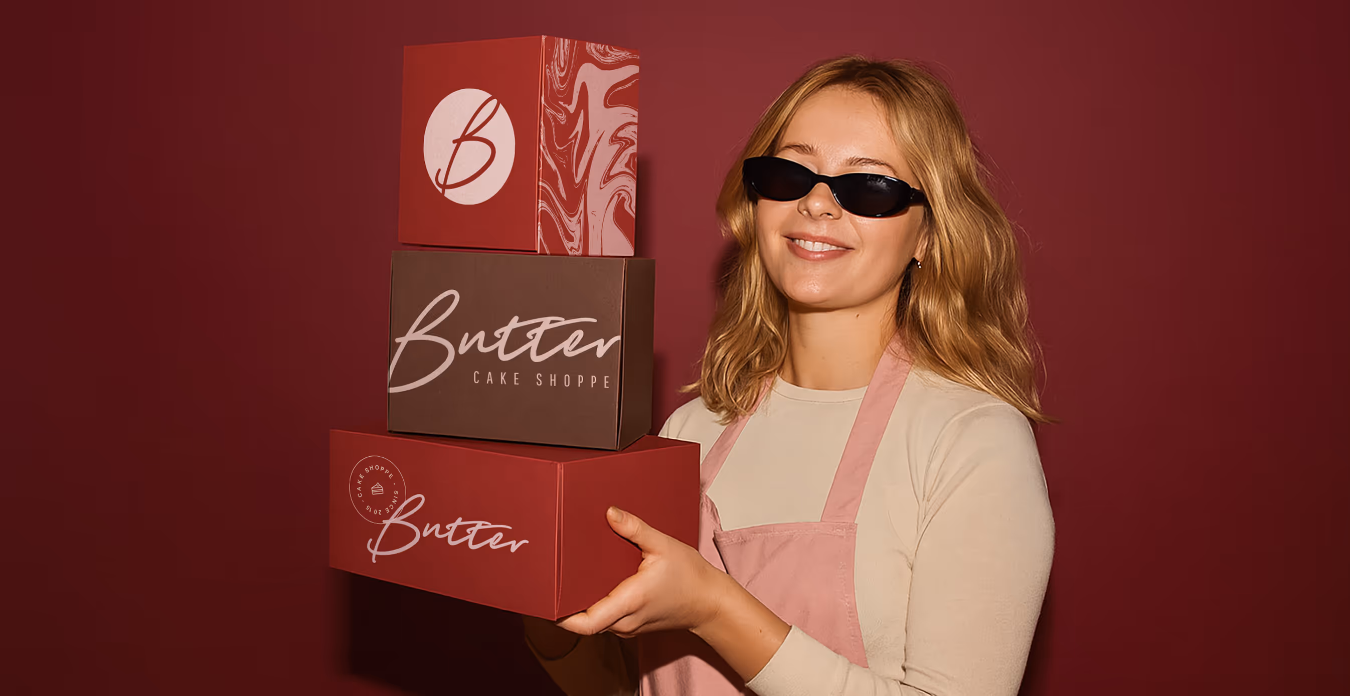 Smiling woman in sunglasses and apron holds three stacked branded boxes with "Butter Cake Shoppe" logo against a burgundy backdrop.