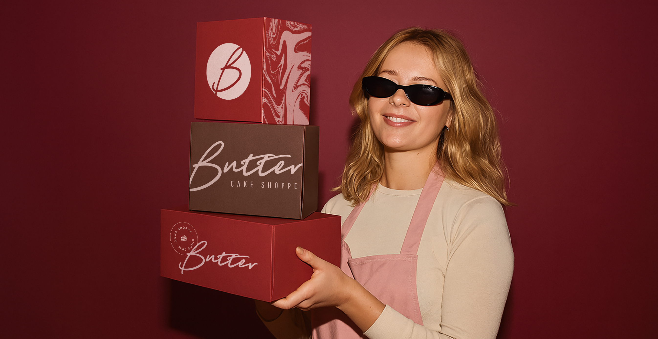 Smiling woman in sunglasses and apron holds three stacked branded boxes with "Butter Cake Shoppe" logo against a burgundy backdrop.