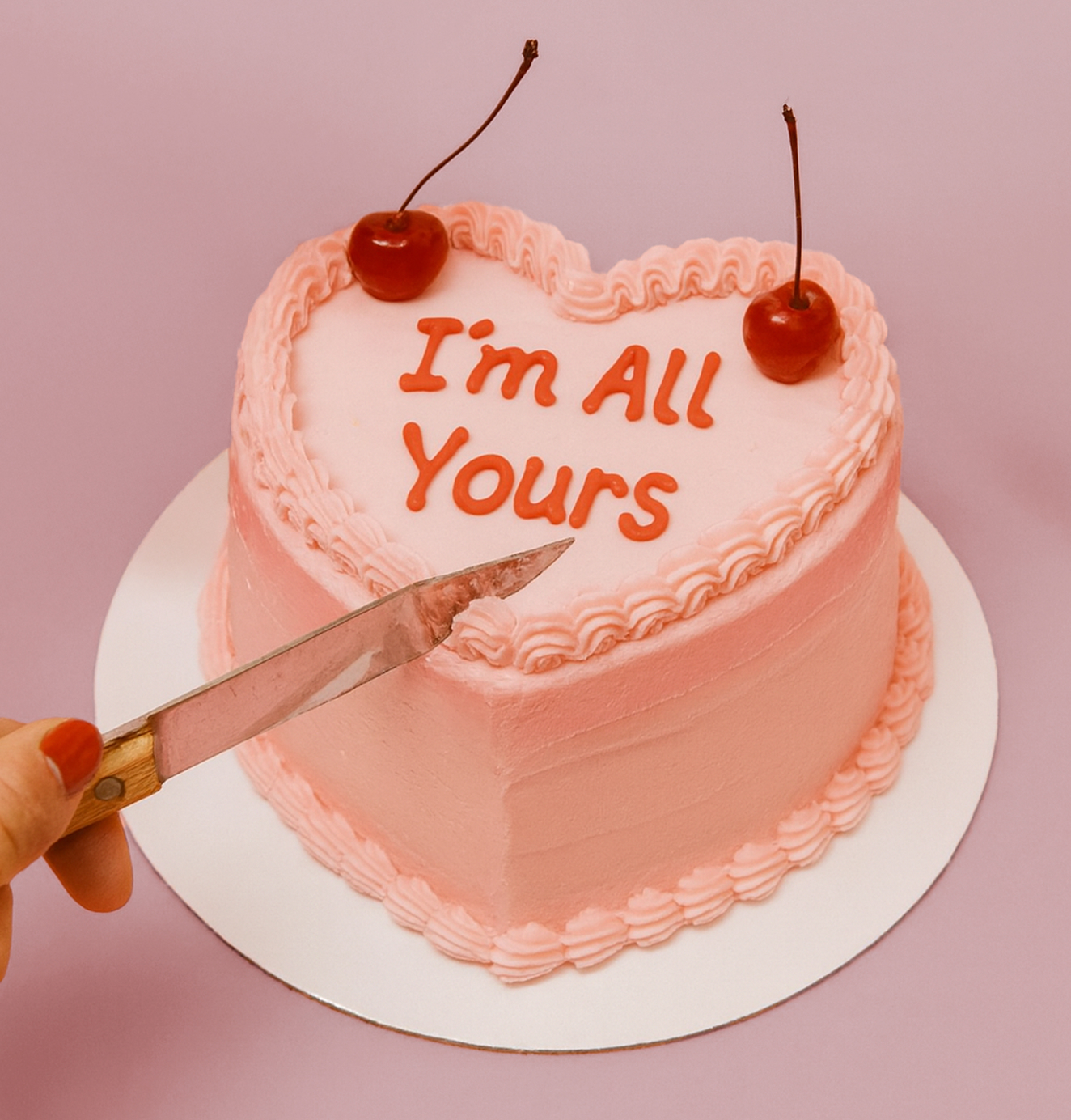 Heart-shaped pink frosted cake with the words “I’m All Yours” in red icing, topped with two cherries, as a hand prepares to cut it.
