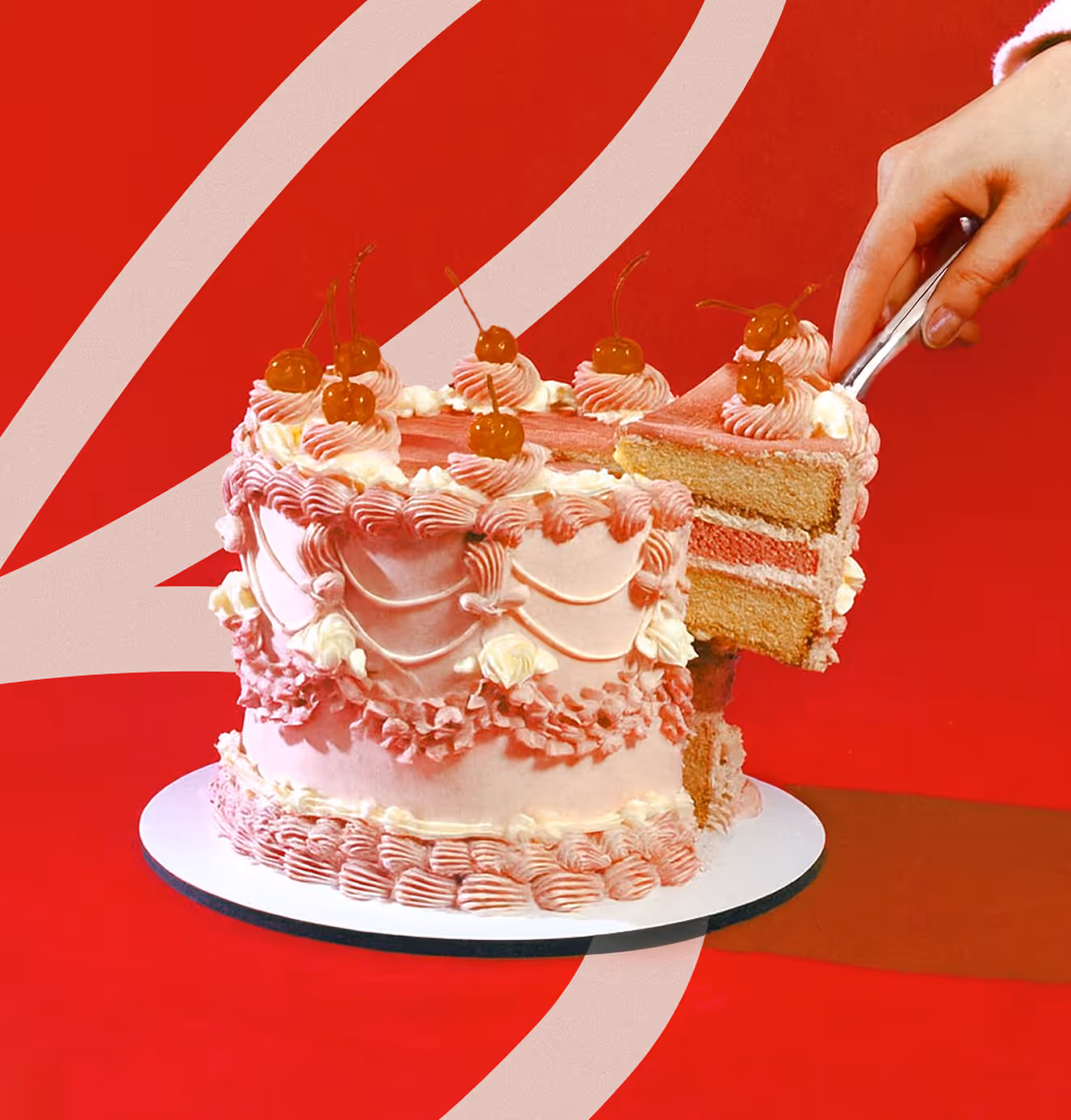 Pink frosted layer cake with cherries and piped decorations as a hand lifts out a slice with a cake server.