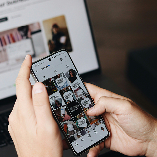 A person holding a smartphone and browsing an Instagram profile grid, with a laptop open in the background.