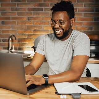 Smiling man wearing glasses and a gray t-shirt working on a laptop at a kitchen table with a notebook and phone beside him.