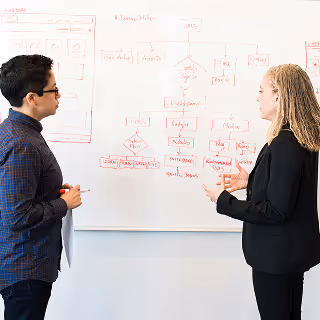 Two professionals standing in front of a whiteboard with a red marker-drawn flowchart, discussing a business or project plan.