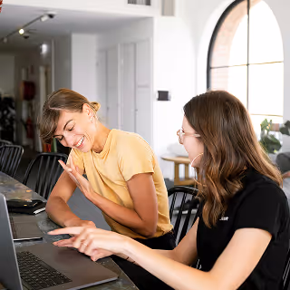 Two women sitting at a counter in a bright, modern workspace, smiling and looking at a laptop while engaging in a lively conversation.