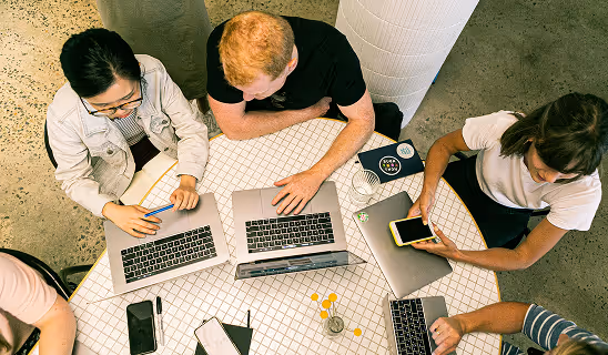 Overhead view of a group of people collaborating at a round table with laptops, notebooks, and a smartphone.