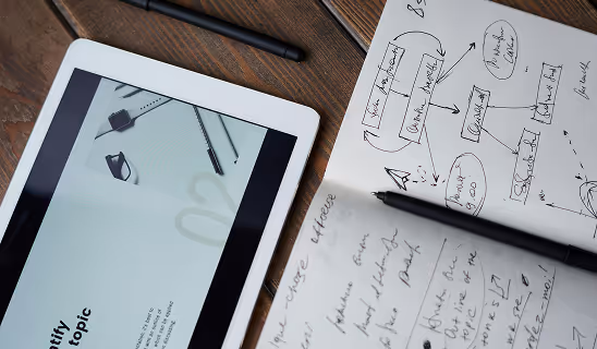 Tablet displaying a sleek presentation next to an open notebook filled with sketched diagrams and handwritten notes, with pens scattered around.