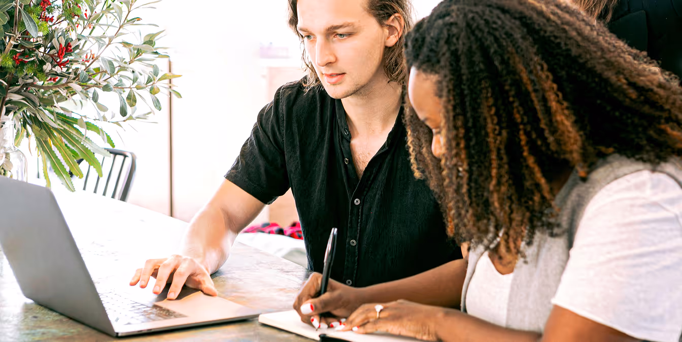 Two colleagues at a table, one working on a laptop and the other taking notes in a notebook, with a vase of greenery in the background.