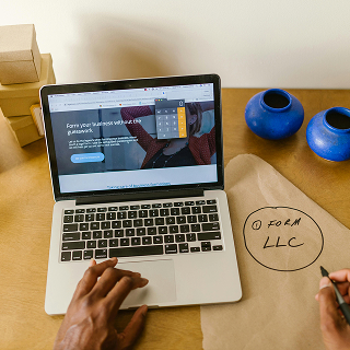 Person working on a laptop showing a business formation website, with a hand writing “Form LLC” on a piece of brown paper beside it.
