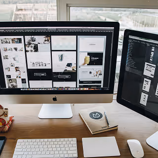 Dual computer monitors on a desk displaying website layout designs, with a notebook, keyboard, and other workspace items nearby.