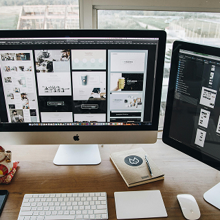 Dual computer monitors on a desk displaying website layout designs, with a notebook, keyboard, and other workspace items nearby.