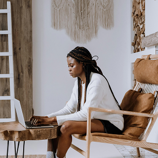 Woman sitting in a modern, bohemian-style living space, working on a laptop at a small wooden table.