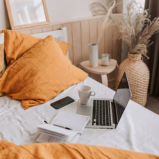 Cozy bed with mustard-colored pillows, an open laptop, a smartphone, a coffee cup, and a notebook with a pen on the duvet.