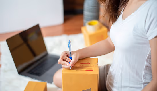 Woman writing on a cardboard shipping box with a pen while sitting near a laptop, preparing an order for delivery.