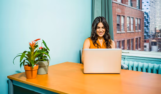 Smiling woman working on a laptop at a desk with potted plants, set against a light blue wall and window view of city buildings.