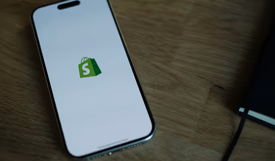 Close-up of a smartphone on a wooden surface displaying the Shopify logo on its screen.