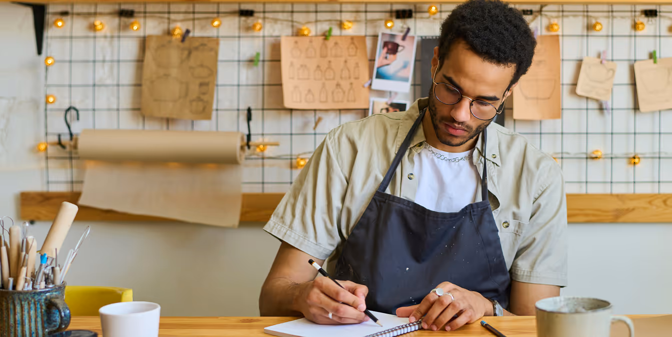 Man wearing glasses and an apron sketching product designs at a workbench, with tools and concept drawings in the background.