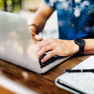 Hands typing on a silver laptop keyboard at a wooden table, with a watch on the left wrist and a notebook in the background.