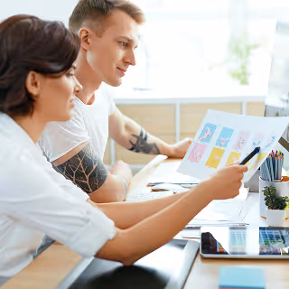 A man and woman at a desk collaborate while reviewing colorful printed design samples, with a computer monitor and graphic tools nearby.