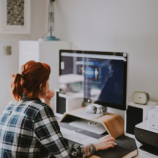 A person with red hair tied back, wearing a plaid shirt, sits at a desk in a home office, focused on a large monitor displaying a design interface.