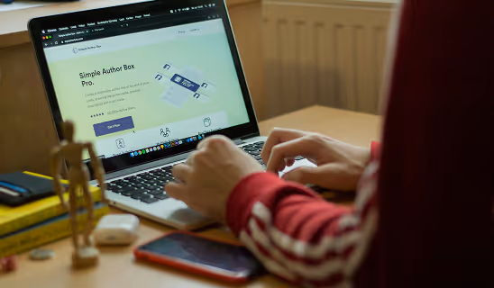 A person types on a MacBook displaying a WordPress plugin product page, seated at a desk with books and a phone nearby.